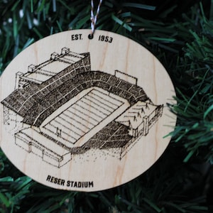 May include: A wooden ornament with a laser-engraved illustration of Reser Stadium, a football stadium. The ornament is round and has the text "EST. 1953" and "RESER STADIUM" on it.