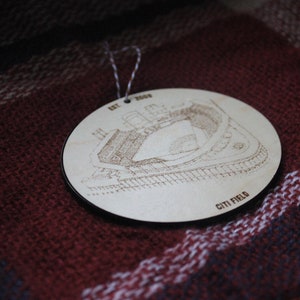 May include: A wooden ornament shaped like Citi Field, a baseball stadium in Queens, New York. The ornament is light brown and features a detailed engraving of the stadium's exterior. The text "EST 2009" and "CITI FIELD" are engraved on the ornament.