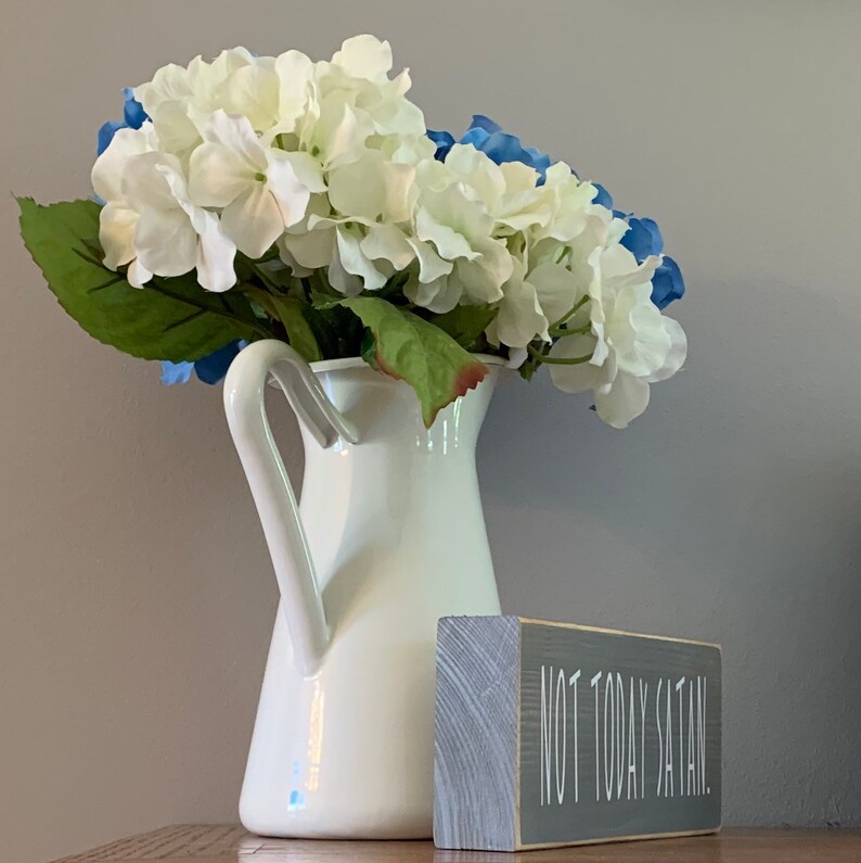 May include: White ceramic pitcher with a handle, filled with white and blue hydrangeas. A gray wooden sign with the text "Not Today Satan." is in the foreground.