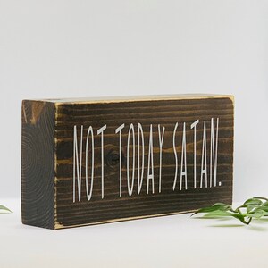 May include: A dark brown wooden block sign with the text "NOT TODAY SATAN." in white lettering.