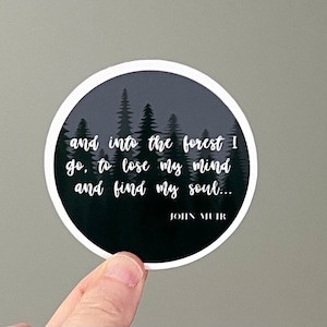 May include: A round sticker with a black and white design. The sticker features a silhouette of a forest with the quote "and into the forest I go, to lose my mind and find my soul..." by John Muir.