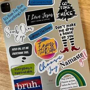May include: A collection of colorful stickers with various phrases and designs, including "Go Take a Hike", "I Love Jesus (But I Cuss a Little)", "Enjoy the Ride", "Choose Joy", and "Namaste Y'all".