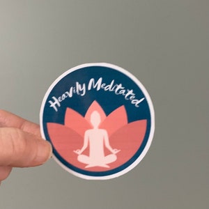 May include: A round sticker with a blue background and a white silhouette of a person meditating in a pink lotus flower. The text "Heavily Meditated" is written in white around the edge of the sticker.