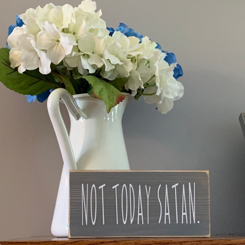 May include: A gray wooden sign with the text "NOT TODAY SATAN." in white lettering. The sign is sitting on a table in front of a white pitcher with white and blue flowers.