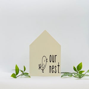 May include: A white wooden house-shaped sign with black text that reads "our nest" and a small black bird illustration.
