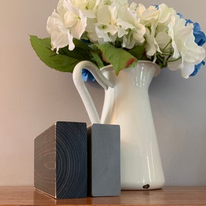 May include: Two wooden blocks, one dark gray and one light gray, are sitting on a wooden surface. The blocks are both rectangular and have a wood grain pattern. A white pitcher with a bouquet of white flowers is in the background.