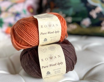 May include: Two balls of Rowan Pure Wool 4ply yarn. One is a burnt orange color, and the other is a dark brown. Each ball has a label with the Rowan logo and product information. The yarn is made of 100% wool.