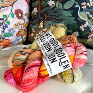 Może przedstawiać: Dwa motki włóczki w jasnym pomarańczowym, żółtym i różowym kolorze. Włóczka jest oznaczona "Woool EooLen BooVon Joon" i "Wicked Pretty Yarn Pretty Yarn For All".
