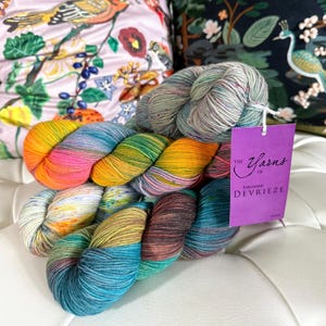May include: Four skeins of colourful yarn in shades of blue, green, orange, pink, and brown. The yarn is arranged in a pile on a white surface. A purple tag with the text "The Yarns of Richard Devrieze" is visible.