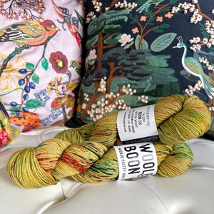 Może przedstawiać: Motka zielonej, żółtej i pomarańczowej przędzy z czerwonymi i niebieskimi plamkami. Przędza jest oznaczona jako "Wool Boon" i "Wicked Pretty Yarn".