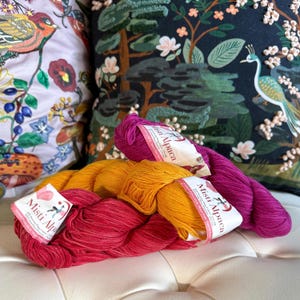 May include: Two skeins of yarn, one orange and one pink, with the label "Misti Alpaca" on each. The yarn is arranged on a white surface.