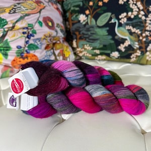Op de afbeelding: Een bol kleurrijk garen met een roze, paarse, blauwe en groene kleurstelling. Het garen is gelabeld met "BASIC DALE" en "SUPERWASH 100% MERINO WOOL".