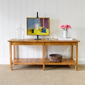 May include: A wooden console table with two shelves, holding a painting, a large glass bottle, a vase of pink flowers, and a woven basket. The painting has a yellow background and blue, white, and purple elements. The table is on a beige rug.