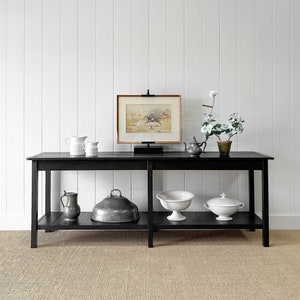 May include: A black console table with a lower shelf displays various decorative items. A framed equestrian print sits on top, alongside white pitchers, a teapot, and a potted plant. The lower shelf holds metal and white ceramic serving pieces.