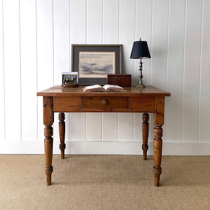 May include: A rectangular wooden desk with four turned legs and a single central drawer featuring a round knob. On the desk surface sits an open book, a framed landscape painting, a small wooden box, and a brass lamp with a black shade.