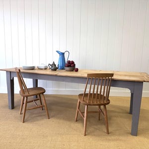 May include: A long, rectangular wooden dining table with a gray base and natural wood top. Two wooden chairs are positioned on either side. A blue pitcher, teapot, and bowl of fruit are on the table. The table is approximately 100 inches long.