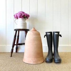 May include: A terracotta-colored, lidded garden pot stands next to a pair of black Hunter rain boots. A small wooden stool holds a white pitcher filled with pink peonies. The scene is set against a white paneled wall.