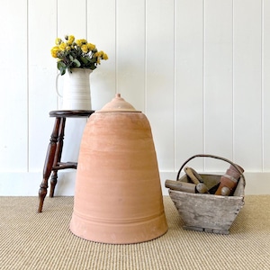 May include: A terracotta pot with a lid, a wooden stool, and a wooden basket with tools. The pot is a light brown color and has a smooth surface. The stool is dark brown and has a simple design. The basket is made of weathered wood and has a handle. The tools in the basket are made of wood and metal.