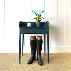 May include: A teal-colored antique desk with a drawer and a small shelf, holding a potted plant with blue flowers and a candle. A pair of black riding boots stand in front of the desk. The desk is approximately 30 inches tall.