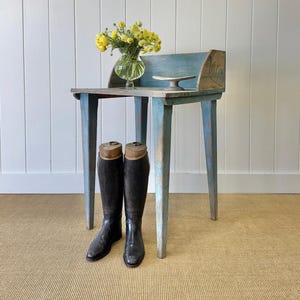 May include: A blue painted wooden table with four legs and a shelf. The table has a worn, distressed look. There are two black leather riding boots with wooden boot trees on the floor in front of the table. A vase with yellow flowers sits on the table.