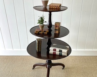 May include: A three-tiered mahogany table with a turned pedestal base and three round shelves. The table is decorated with small potted plants and books.