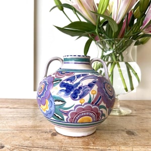 May include: A ceramic vase with two handles, featuring a colorful floral and bird design in shades of blue, purple, and yellow. The vase is set on a wooden surface, with a glass vase of flowers in the background.