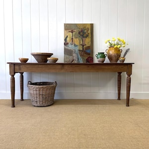May include: A long, dark wood table with turned legs, displaying a variety of decorative items. A woven basket sits below. The table holds wooden bowls, vases with flowers, and a framed painting. The room has white paneling and a neutral carpet.