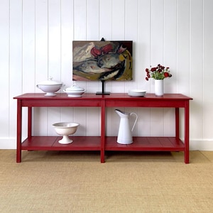 May include: A red wooden console table with two shelves. The table has a white pitcher, white bowls, and white plates on top. There is a painting of fish on the wall behind the table.