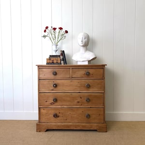 May include: A light brown wooden dresser with five drawers and round wooden knobs. A white bust, books, and a vase of red flowers sit on top. The dresser is against a white paneled wall.