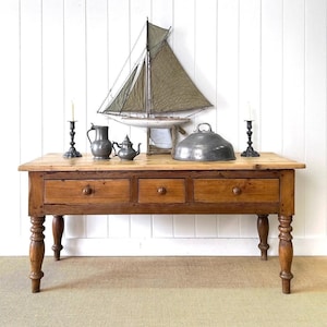 May include: A large, antique wooden table with three drawers and turned legs. The tabletop is light-colored wood, and the drawers are a darker shade. Decorative items include a model sailboat, pewter pitcher, teapot, and candle holders.