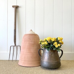 May include: A terracotta pot with a white top, a vintage metal pitcher with two handles, and a yellow flower arrangement. The items are on a brown woven rug in front of a white wood wall.