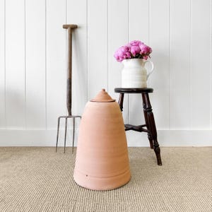 May include: A terracotta cloches with a wooden handle, a white pitcher with pink flowers, and a wooden stool. The items are on a light brown rug.