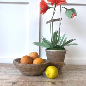 May include: A rustic wooden bowl with a handle containing three oranges, placed next to a single yellow lemon and a terracotta pot holding three orange paper poppy flowers with green leaves and a small rectangular tag attached to a stem.