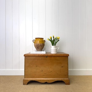 May include: A wooden chest with a curved base, topped with a yellow and brown ceramic vase, a stack of books, and a white pitcher holding yellow tulips. The chest is made of light-colored wood and sits on a beige carpet.