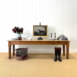 May include: A long, wooden dining table with turned legs, set with various decorative items. A framed painting of a horse, candles, and a metal cloche are on the table. A basket with a plaid blanket and riding boots are on the floor.