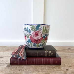 May include: A white ceramic planter with a hand-painted floral design in red, blue, and green. The planter sits atop two antique books with a decorative tassel. The planter has a blue rim and a floral pattern.
