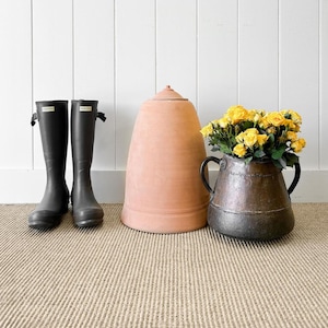 May include: A terracotta pot with a rounded top, a black metal pot with handles, and a bouquet of yellow flowers. Two black rain boots are on the floor in front of the pots.