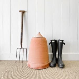 May include: A terracotta pot with a lid, a garden fork, and a pair of black rain boots on a brown rug.