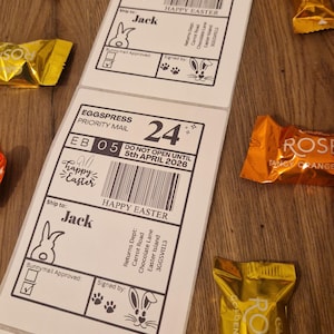 Peut inclure: Étiquettes d'expédition blanches sur le thème de Pâques avec le texte "Happy Easter" et une illustration de lapin. Les étiquettes sont entourées de bonbons emballés individuellement dans du papier d'aluminium doré et orange.