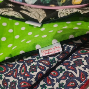 May include: A stack of fabric squares with different patterns. The top square is black with a white design. The second square is green with white polka dots. The third square is white with a blue and red paisley pattern. The bottom square has a red border. A white label with the text "Desert Pea Industries" is attached to the green square.