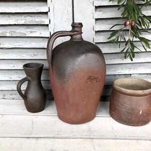 May include: Three brown ceramic pottery jugs of different sizes and shapes. The largest jug has a wide mouth and a handle. The smallest jug has a narrow mouth and a handle. The middle jug has a wide mouth and no handle.