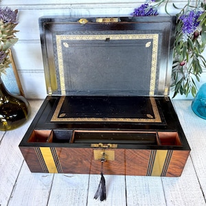 Puede incluir: Un escritorio antiguo de madera abierto con interior negro y ribetes dorados. El exterior es de madera oscura con detalles de latón y una cerradura con borla. La caja está sobre una superficie de madera blanca.