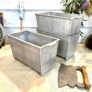 Op de afbeelding: Een stapel van drie rechthoekige, zilverkleurige metalen broodvormen met een vintage uitstraling. De vormen hebben een licht getextureerd oppervlak en kleine handgrepen aan de zijkanten. Een antieke strijkijzer met houten handvat is ook zichtbaar.