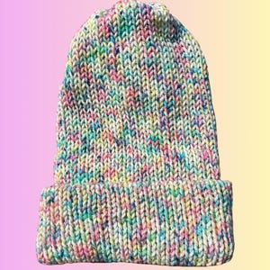 May include: A hand-knit beanie hat featuring a multi-colored speckled yarn pattern. The knit fabric displays a vibrant mix of pink, blue, yellow, and green flecks against a cream-colored base. The hat includes a wide, folded cuff at the bottom.