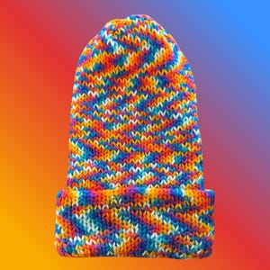 May include: A knit beanie hat featuring a vibrant, multi-colored variegated yarn pattern. The design incorporates a blend of bright orange, yellow, blue, purple, and white fibers throughout the fabric, finished with a wide, folded cuff at the base.