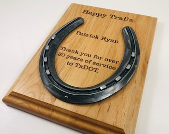 May include: A wooden plaque with a horseshoe attached to it. The plaque is engraved with the text "Happy Trails", "Patrick Ryan", "Thank you for over 30 years of service to TxDOT."
