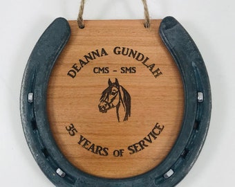 May include: A wooden plaque with a horseshoe frame, engraved with the text "Deanna Gundlah CMS - SMS 35 Years of Service". A horse's head is also engraved on the plaque.