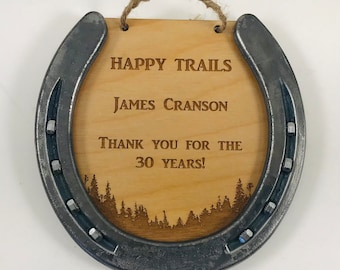May include: A wooden horseshoe with a metal frame, engraved with the text "Happy Trails", "James Cranson", and "Thank you for the 30 years!" The horseshoe is hanging from a twine rope.
