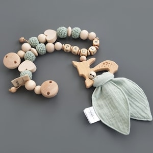 Op de afbeelding: Een houten baby-bijtje met een hertenbedel, een groen stoffen label en een houten kralenketting met groene en witte kralen. De ketting heeft houten kralen met de naam "PAULA" erop.