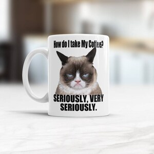 Puede incluir: Taza de café de cerámica blanca con una ilustración de un gato gruñón y el texto "¿Cómo tomo mi café? En serio, muy en serio."
